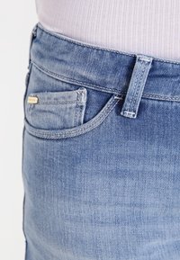 Jasnoniebieskie dżinsy z denimu o wąskim kroju. Posiadają małą przednią kieszeń, przeszycia dekoracyjne oraz metalowy detal na pasku.