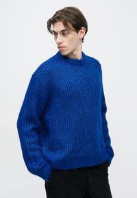 Kék kötött pulóver texturált felülettel, kerek nyakkivágással, széles ujjakkal és bordázott mandzsettákkal. Sötét nadrág felett viselve, egyszerű háttér előtt.