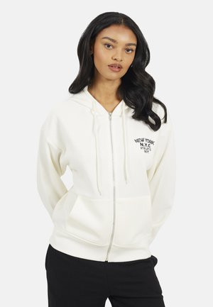 Donna con lunghi capelli scuri che indossa una felpa bianca con zip e la scritta "New York N.Y.C Athletic Dept", abbinata a pantaloni neri, in piedi su sfondo bianco.