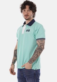 Homme aux cheveux bouclés, portant un polo vert menthe avec un col et un logo bleu marine, un jean bleu, et des tatouages détaillés sur les deux avant-bras.