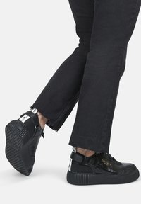Schwarze Sneakers mit glänzender Oberfläche, strukturierten Details und einer klobigen Sohle. Getragen mit verkürzten schwarzen Jeans mit fransigen Kanten.