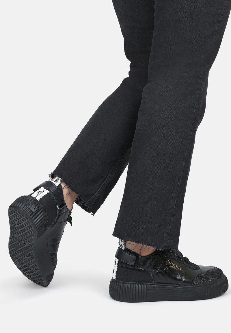 Schwarze Sneakers mit glänzender Oberfläche, strukturierten Details und einer klobigen Sohle. Getragen mit verkürzten schwarzen Jeans mit fransigen Kanten.
