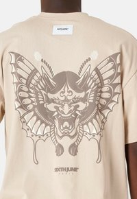 Camiseta de algodón beige con un gran gráfico gris de un feroz dragón con alas de mariposa en la parte posterior, y etiqueta de la marca en el cuello.