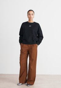 Felpa nera con logo bianco, abbinata a pantaloni ampi marroni. Il modello indossa scarpe argentate lucide. Sfondo minimalista.