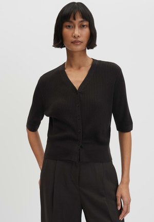 Frau mit kurzen schwarzen Haaren, die eine schwarze gerippte, geknöpfte Strickjacke mit ellbogenlangen Ärmeln und schwarze Hose trägt, steht vor einem einfarbigen Hintergrund.