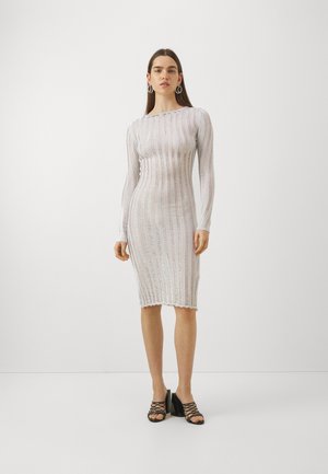 Robe bodycon en tricot blanche transparente avec une texture à côtes verticales. Manches longues et ourlet en forme de vague. Associée à des sandales à talons carrés noires à brides.