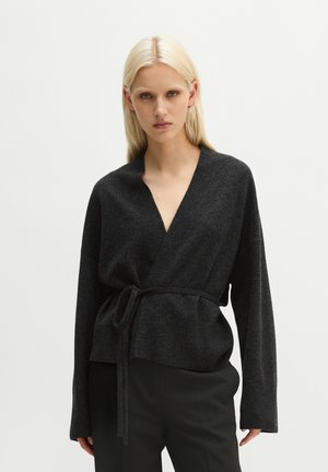 Donna con capelli biondi lisci che indossa un maglione avvolgente grigio scuro legato in vita e pantaloni neri, in piedi davanti a uno sfondo chiaro e uniforme.
