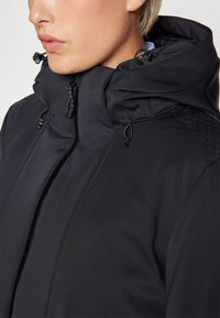 Must must jacket, mille kõrge krae, reguleeritava kapuutsiga, millel on nööbid, ja tekstuuriga muster ühel õlal. Sile kangas.