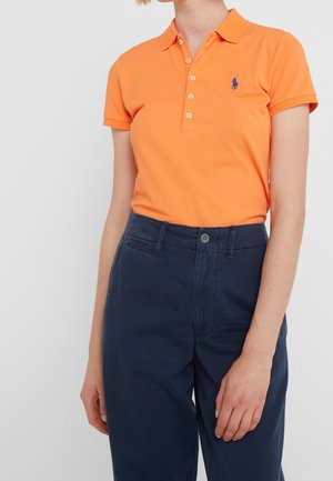 Pomarańczowa bawełniana koszulka polo z klasycznym kołnierzykiem, z małym niebieskim haftowanym logo na piersi, w zestawie z granatowymi spodniami.