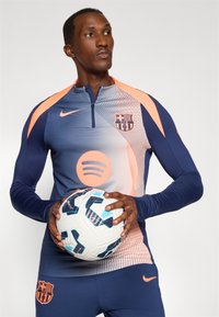 Haut d'entraînement à manches longues FC Barcelone de Nike en bleu et rose, avec un col zippé, le logo du club et un graphique Spotify, accompagné d'un ballon de football.