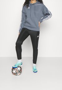 Un sweat à capuche gris avec des rayures blanches, un pantalon de sport noir et des chaussures de football colorées. Un ballon de football est placé aux pieds.