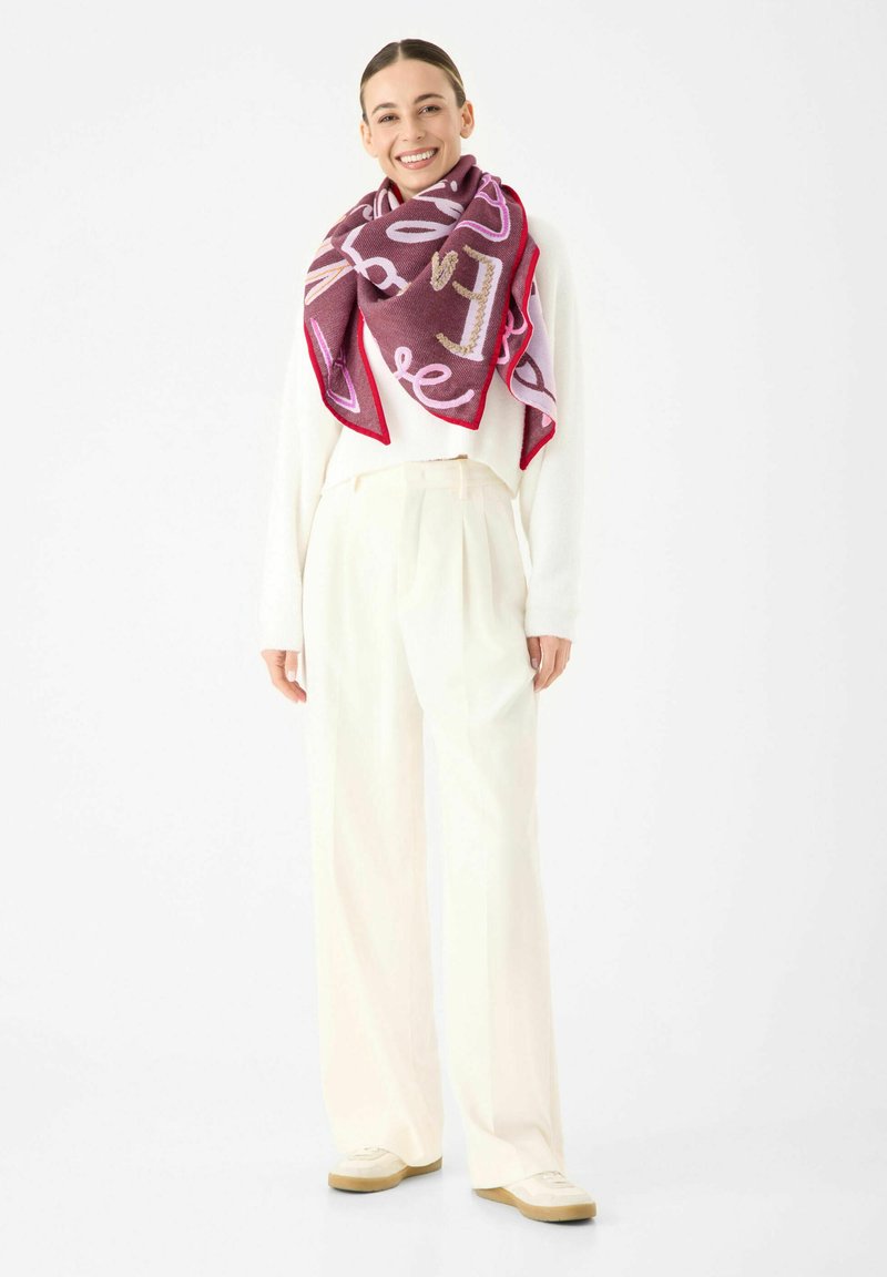 Lachende vrouw met een witte trui en broek, en een grote lila en roze sjaal met patroon, staand tegen een witte achtergrond.