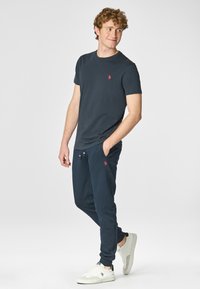 Maglietta navy con logo rosso, abbinata a pantaloni joggers coordinati, presenta vita con cordino e tasche laterali. Il modello indossa sneakers bianche.