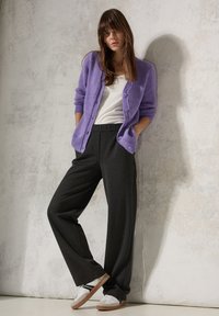 Lila fluffig cardigan över vit skjorta, kombinerat med mörkgrå vida byxor och vita sneakers med bruna sulor, mot en texturerad bakgrund.