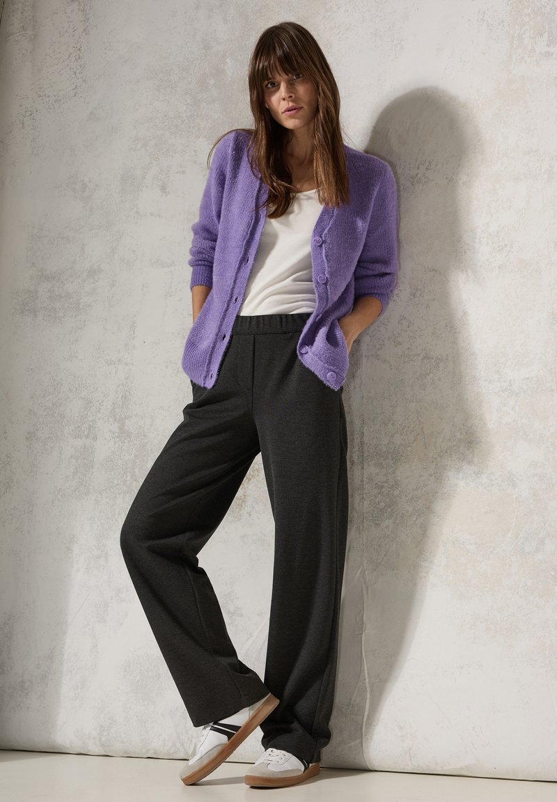 Lila fluffig cardigan över vit skjorta, kombinerat med mörkgrå vida byxor och vita sneakers med bruna sulor, mot en texturerad bakgrund.