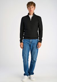 Maglione nero in maglia con colletto a zip quarti, abbinato a jeans in denim blu e sneakers bianche. Design semplice e casual; tessuto texturizzato.