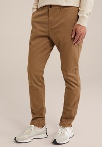 Hnědé bavlněné chino kalhoty se slim fit střihem, předními kapsami a zapínáním na knoflík, doplněné béžovými a šedými teniskami s bílými tkaničkami.