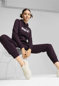 Completo di felpa viola Puma e pantaloni della tuta con logo bianco, materiale morbido, vestibilità comoda, abbinato a sneaker beige chiaro su una sedia.