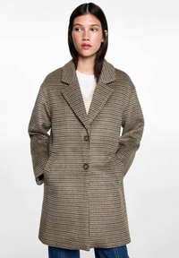 Brun houndsstooth frakke med slagkrave, to forlommer, tre knapper og en løs pasform. Stoffet har en struktureret overflade.