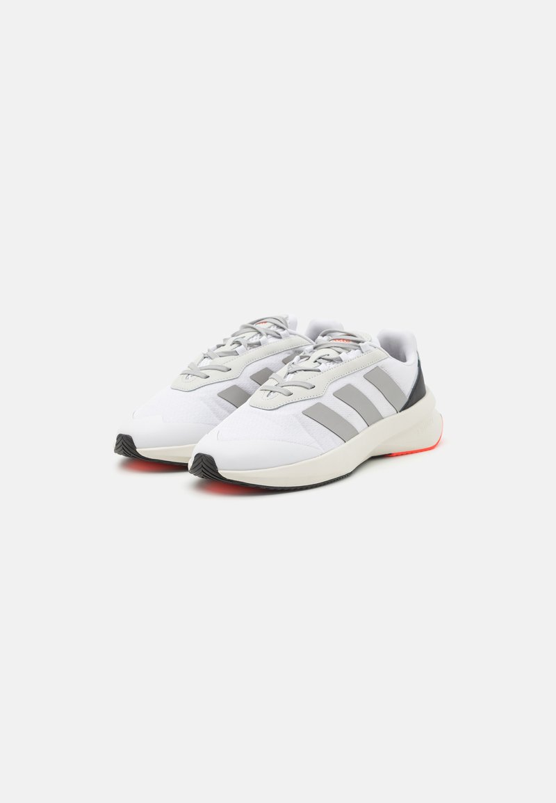 Scarpe sportive bianche con accenti grigi, tessuto texturizzato, forma arrotondata e suola nera. Presentano strisce grigie sui lati e dettagli rossi sulla suola.