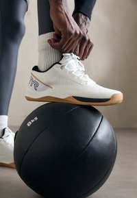 Persona, kura sapina kurpes baltās sporta kurpes ar brūniem zole, kāja atpūšas uz 8 kg melnā medicīnas bumbas uz pelēka grīdas.