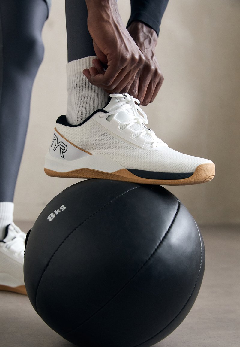 Persona, kura sapina kurpes baltās sporta kurpes ar brūniem zole, kāja atpūšas uz 8 kg melnā medicīnas bumbas uz pelēka grīdas.