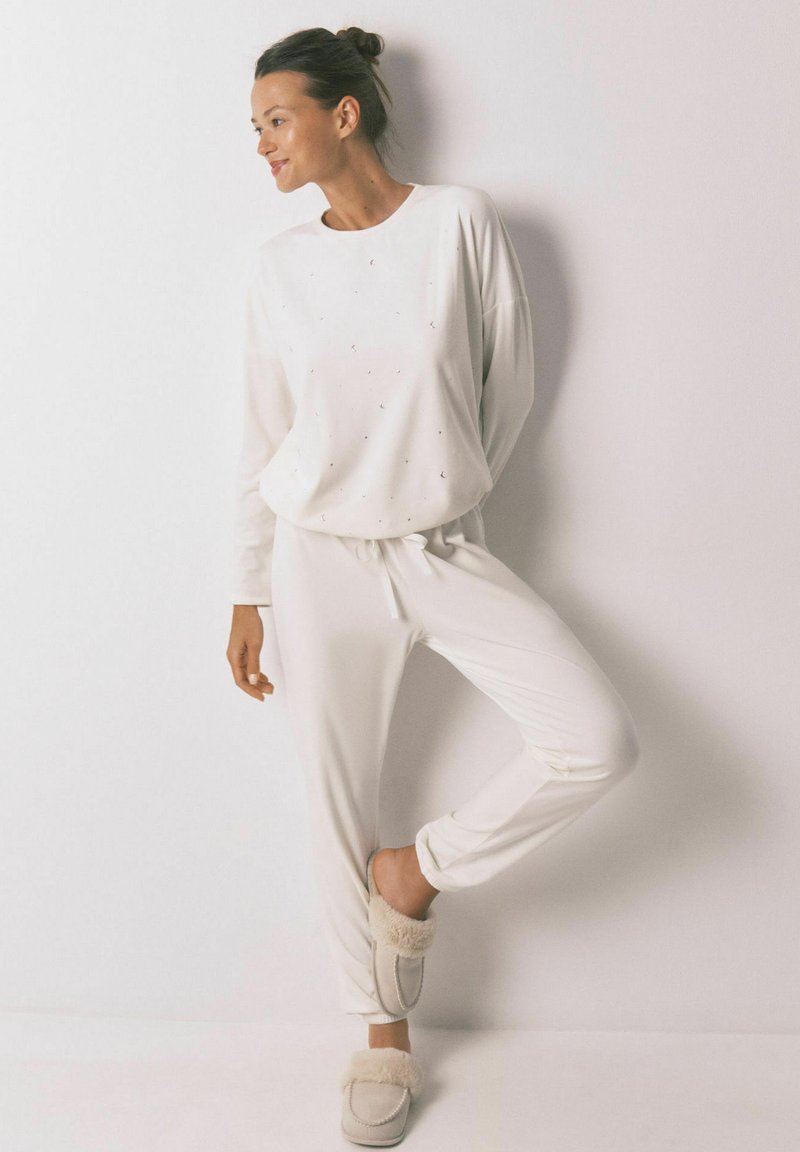 Hvidt loungewear-sæt bestående af en langærmet top med små, subtile udsmykninger og tætsiddende jogger-bukser, suppleret med beige slippers.