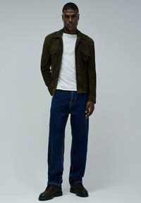 Chaqueta de ante marrón con bolsillos, camiseta blanca, jeans azules y zapatos negros con cordones. El modelo se presenta con confianza frente a un fondo gris claro.