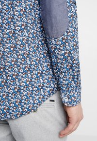 Manche de chemise avec un motif floral bleu, blanc et marron, arborant un patch de coudière bleu uni et des boutons blancs sur le poignet, portée avec un pantalon gris clair.