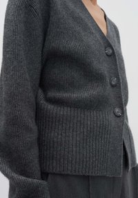 Gros plan sur une personne portant un cardigan en maille côtelée gris foncé avec de gros boutons et un pantalon foncé assorti.