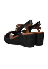 Zwarte wedge sandalen met een geweven plateauzool, verstelbare enkelbandjes en een gouden gesp, in een hoek gepositioneerd om de achterkant en zijkant te tonen.
