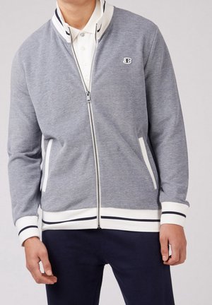 Homme portant une veste zippée gris clair avec des poignets et un col blancs, superposée à une chemise blanche boutonnée, et un pantalon sombre.