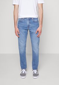 Jeans en denim bleu clair avec une coupe droite, cinq poches et des coutures visibles, associés à des baskets grises et un t-shirt blanc uni.