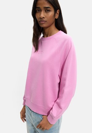 Kobieta o długich ciemnych włosach, ubrana w luźną różową bluzę i jasnoniebieskie dżinsy, stojąca na jednolitym białym tle.