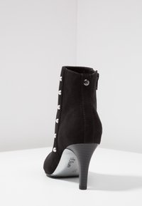 Black suede ankle boot with a pointed toe, featuring a side zipper and decorative metallic buttons along the outer side. Stiletto heel.