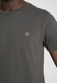 Šedé tričko s krátkým rukávem vyrobené z měkké bavlny. Na levé straně hrudi je malé bílé logo. Má přiléhavý výstřih a hladkou texturu.