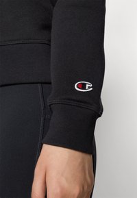 Črna majica s kapuco z rebrastimi manšetami; izvezen logotip na rokavu. Tekstura je gladka, oblikovanje je tesno prilegajoče.