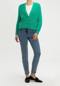 Zelený žebrovaný cardigan s V-výstřihem a zapínáním na knoflíky, v kombinaci s světle modrými džíny a černými botami, na neutrálním pozadí.