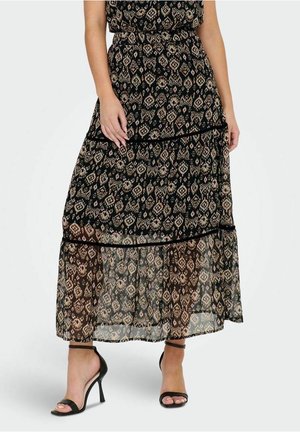 Kvinde iført en sort maxinederdel med beige geometriske mønstre og vandrette sømdetaljer, kombineret med sorte højhælede sandaler med ankelstrop.