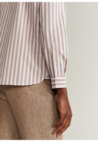 Camisa de manga larga con rayas rosa claro y blancas, con puños abotonados, mostrada sobre una tela texturizada de color marrón claro.