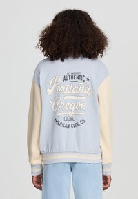 Jasnoniebieska kurtka zapinana na zamek z kremowymi rękawami, z grafiką "Portland Oregon" w białym kolorze, żeberkowane mankiety i dół. Miękka tekstura materiału.