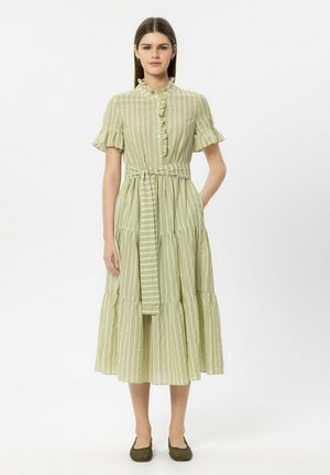 Frau steht und trägt ein hellgrünes, vertikal gestreiftes Midikleid mit kurzen, gerafften Ärmeln, einem gerafften Kragen und einem gebundenen Taillengürtel.