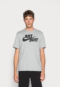 Pilkas medvilnės marškinėlis su juodu užrašu "JUST DO IT" ir Nike swoosh grafika, apvaliu kaklu, trumpomis rankovėmis ir laisvu kirpimu.