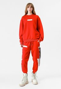 Sudadera roja de gran tamaño con una etiqueta blanca y joggers a juego con acentos estampados, combinados con botas blancas con cordones.