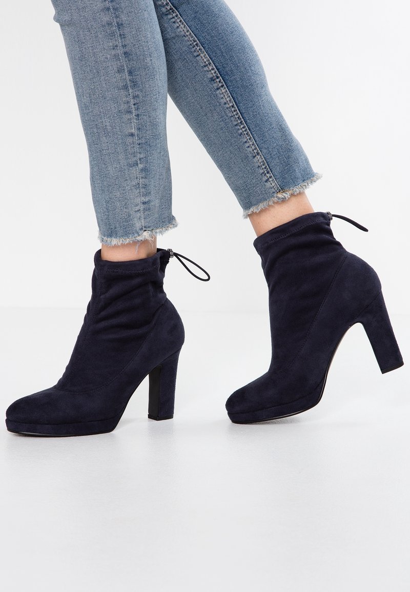 Personne portant un jean bleu clair à ourlet effiloché et des bottines à talons hauts en daim bleu marine avec un détail à cordon, debout sur un sol blanc.