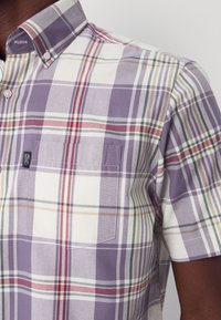 Lähivõte lühikeste varrukatega lilla, valge, punase, rohelise ja beežikarva ruudulise triiksärgi nööpidega kraega ja rinnataskuga tumedanahalise inimese seljas.