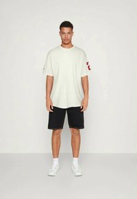 Homem em pé, vestindo uma t-shirt oversized branca com o número 23 em vermelho na manga, calções pretos, meias brancas e ténis brancos, contra um fundo simples.
