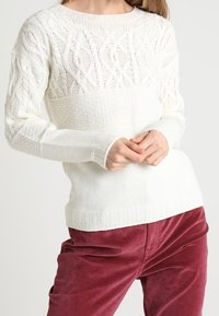 Femme portant un pull blanc en maille torsadée avec des motifs texturés et un pantalon en velours côtelé bordeaux, posant les mains jointes devant elle.