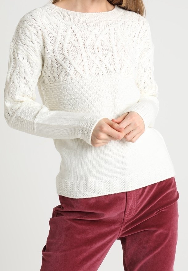 Femme portant un pull blanc en maille torsadée avec des motifs texturés et un pantalon en velours côtelé bordeaux, posant les mains jointes devant elle.