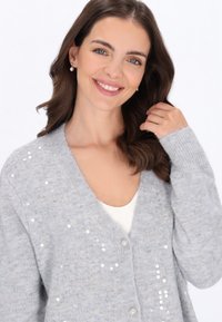 Femeie zâmbitoare cu părul ondulat castaniu, purtând cercei cu perle și un cardigan gri deschis cu mărgele peste un top alb.